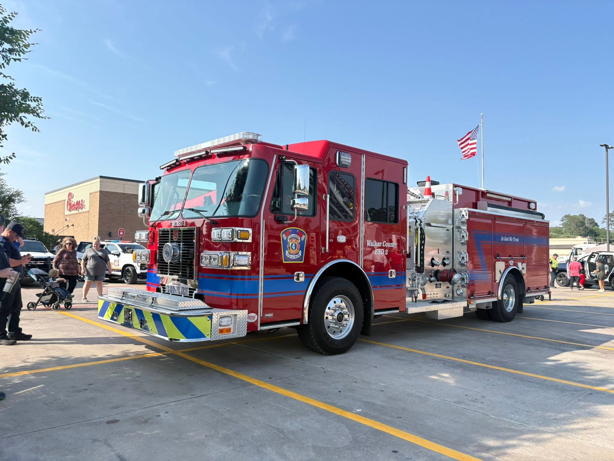 A bright red fire truck is parked in a shopping area, with people engaging nearby, showcasing local emergency services at a community event.